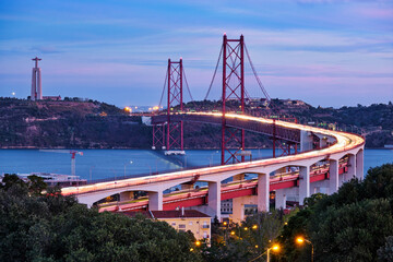View of Lisbon view from Miradouro do Bairro do Alvito viewpoint of Tagus river, traffic with light trails on 25th of April Bridge, and Christ the King statue in the evening twilight. Lisbon, Portugal