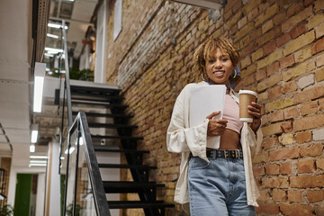 happy african american manager with braces holding papers, startup project, standing in coworking