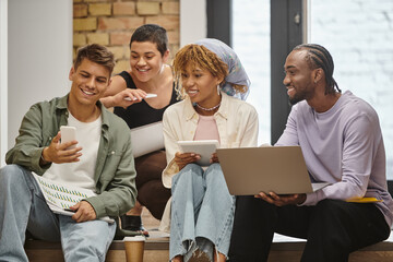 happy diverse team looking at smartphone, holding gadgets, sitting on stairs in coworking, startup