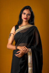 Portrait of a young beautiful girl wearing traditional black saree posing on a brown background.