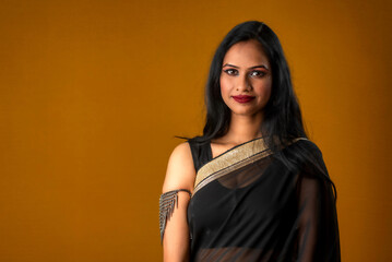 Portrait of a young beautiful girl wearing traditional black saree posing on a brown background.