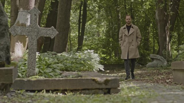 Young Caucasian man in camel trench coat strolling along cemetery at daytime