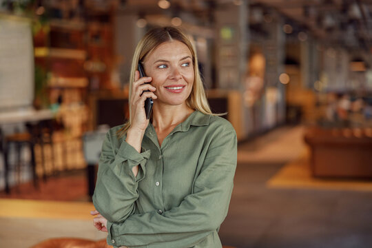Smiling businesswoman is talking phone with client while standing on modern coworking background