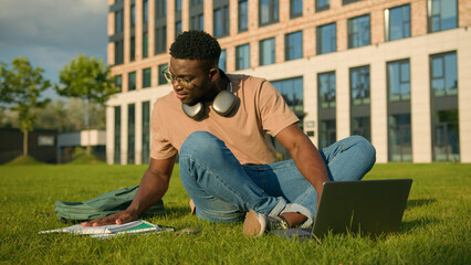 Happy smiling African American man university college academy student guy teenager male studying homework online on grass meadow in park study learning with book copybook laptop prepare for exam study © Yuliia