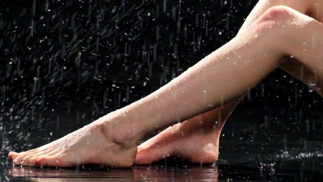 Water drops fall on woman legs. Stream of rain descends on feet of young lady with nude nails on blurry background in studio slow motion