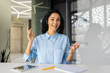 Webcam view, business woman looking at camera with headset talking to customers on video call, Hispanic woman inside office at workplace, advising consumers buyers online support service.
