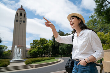 happy asian chinese woman traveler finger pointing into distance at landmark hoover tower under sunny blue sky while touring school campus in California usa at springtime