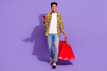 Full length portrait of handsome guy hold mall bags walking use smart phone isolated on purple color background