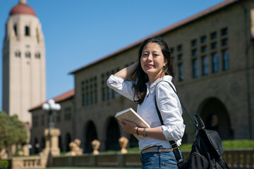 attractive asian Chinese female exchange student holding book and pushing hair back while looking...
