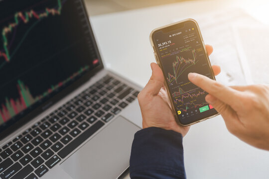 Trader, investor or businessman is pointing on smartphone, laptop on trading stock market graph, candlestick chart as Bitcoin Cryptocurrency price graph chart on screen. Investing money trader concept