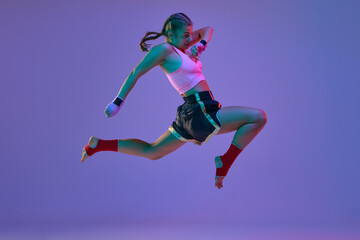 Fototapeta na wymiar Kick in a jump. Teen girl, mma athlete in motion, training against purple studio background in neon lights. Concept of mixed martial arts, sport, hobby, competition, athleticism, strength, ad