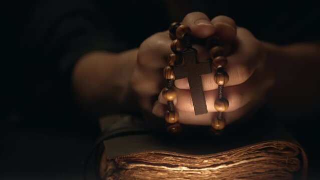 Hands folded in prayer on a Holy Bible in church. Woman praying with a wooden cross. Spirituality religion and hope concept. Supplication believe and faith. Abstract vintage religious dark background