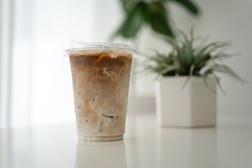An iced milk coffee in plastic cup is placed on white table with white curtain and brightness light as background. Drink and beverage object photo.