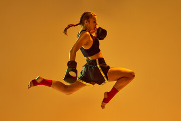 Fototapeta na wymiar Kick in a jump. Teen girl, mma athlete in motion, training against orange studio background in neon lights. Concept of mixed martial arts, sport, hobby, competition, athleticism, strength, ad
