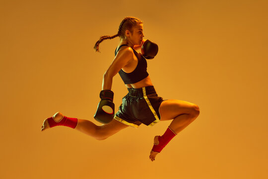 Kick in a jump. Teen girl, mma athlete in motion, training against orange studio background in neon lights. Concept of mixed martial arts, sport, hobby, competition, athleticism, strength, ad