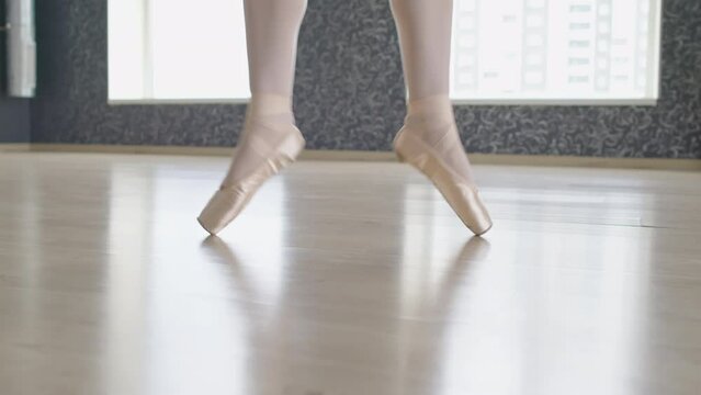 Close up of legs of dancer in pointe shoes with satin ribbon doing ballet poses on tiptoes on floor in light studio