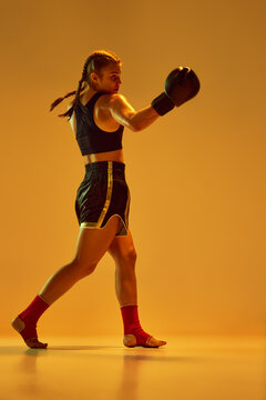 Athletic, serious and concentrated teen girl, MMA fighter training, fighting against orange studio background in neon lights. Concept of mixed martial arts, sport, hobby, competition, strength, ad