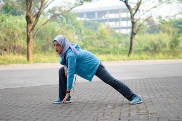 Beautiful Muslim woman working out outdoors in the morning. Sporty woman with a hijab warming up by doing exercises.