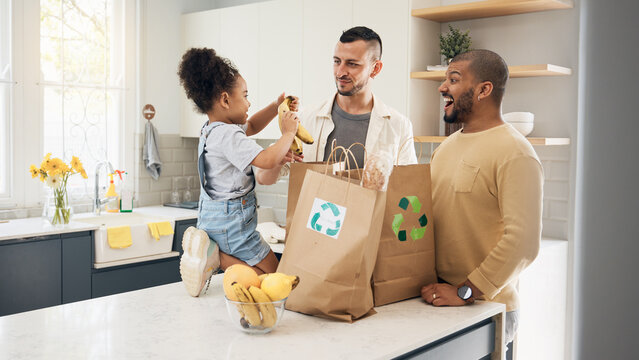 Recycling, blended family or adoption with a girl and gay couple in the home kitchen for sustainability. LGBT, eco friendly or waste with a daughter and parents unpacking bananas from a grocery bag - Powered by Adobe