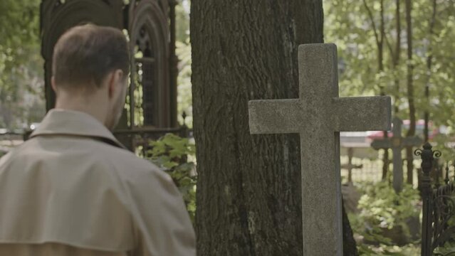 Back view medium shot of man in camel trench coat standing by cross headstone in quiet cemetery at daytime