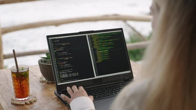 Female soft dev with laptop on remote work day in tropical cafe. Software developer writes program code on computer screen in coworking restaurant with sea view on sunset. Woman coding by ocean.