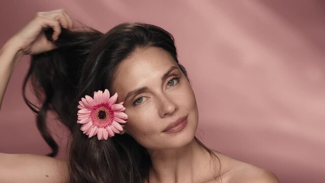 The woman demonstrates healthy, silky hair, throwing it over one shoulder. Portrait of a seminude woman with a gerbera flower in her hair in the studio on a pink background.
