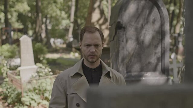 Medium shot of sorrowful grieving man standing in front of headstone at quiet cemetery