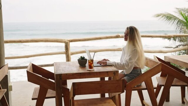 Woman working with laptop in cafe with ocean view, palm trees. Travel blogger typing on computer keyboard sitting in luxury restaurant. SMM manager on remote work day. Female drinks ice coffee by sea.