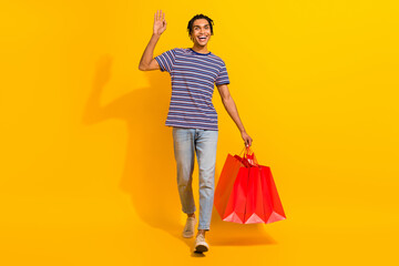 Full size photo of handsome friendly man wear stripped t-shirt go shopping hold new outfit waving palm isolated on yellow color background