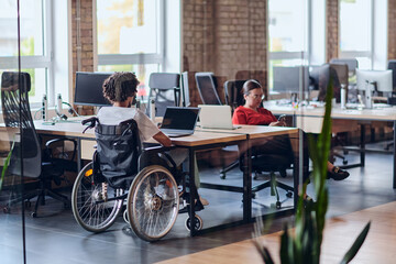 Business colleagues work within a modern startup coworking office, with one of them, an African America in a wheelchair, actively engaging with computers and laptops to collaboratively address various