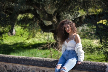 Young, beautiful, brunette woman with curly hair, white top and jeans, sitting on a stone wall, looking at the camera self-confident. Concept beauty, fashion, strength, confidence.