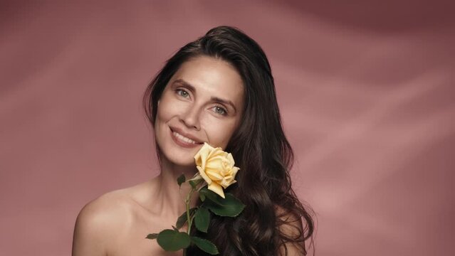 A seminude woman holds a yellow rose on a long stem and inhales its fragrance. Smiling woman holds a rosebud across her face in a studio on a pink background. Spring and summer inspiration.