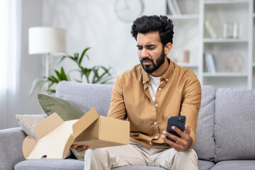 Upset and annoyed man received a parcel, Latin American dissatisfied with the delivery service sits on the sofa in the living room, writes a negative review on the phone.