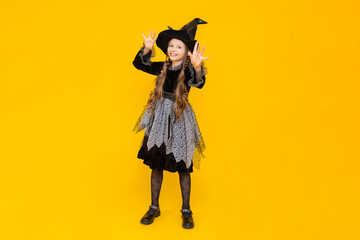 A child in a witch costume. Carnival for children on autumn Halloween. A little girl in a pointed hat and dress is smiling broadly. Happy holiday. Bright yellow isolated background.