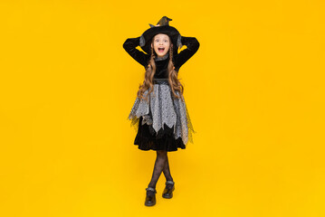 Happy smiling baby on Halloween. A full-length schoolgirl in a carnival witch costume. A young girl holds her hands on a pointed hat. A little enchantress girl on a bright orange isolated background.