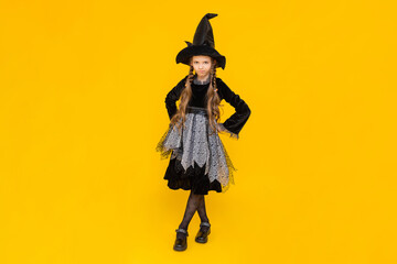 A child in a witch costume. Carnival for children on autumn Halloween. A little girl in a pointed hat shows portrays a harmful, dissatisfied witch. Happy holiday. Bright yellow isolated background.