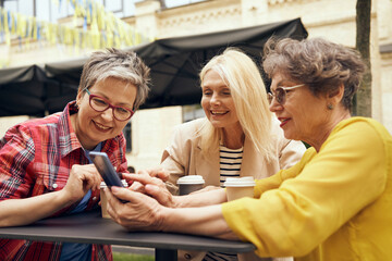 Smiling mature ladies sitting at table with coffee and laughing while watching video on smartphone