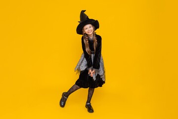 The little witch. A full-length girl in a carnival costume for autumn Halloween. Happy holiday.  A charming young schoolgirl in a pointed black hat and dress. Yellow isolated background.