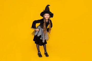 The little witch. A full-length girl in a carnival costume for autumn Halloween. Happy holiday.  A charming young schoolgirl in a pointed black hat and dress. Yellow isolated background.