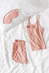 Pink woman pajamas and sleep eye mask on bed. Top view summer pyjama for sleeping. Aesthetic lifestyle flat lay photo, singlet and shorts peach colored, comfort ans stylish wear or clothes