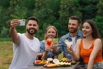 Happy friends with glasses of cocktails taking selfie at table outdoors