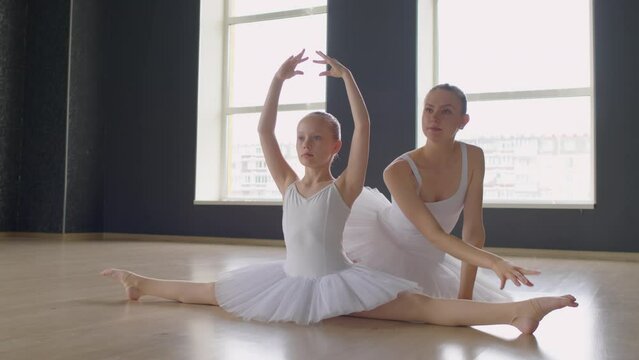 Long shot of little ballerina in white clothes sitting in split and doing exercises with hands above her, teacher sitting behind