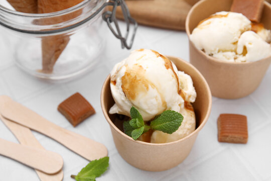 Scoops of ice cream with caramel sauce, mint leaves and candies on white tiled table, closeup