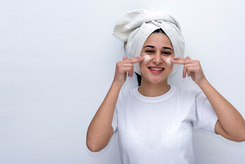 young woman putting cream on her face and happy doing cosmetic treatment on her skin with white towel on her head