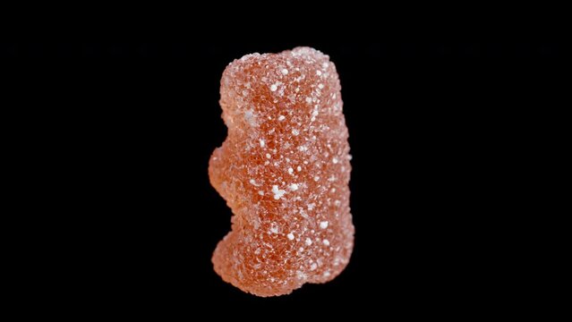 A gummy candy in the shape of a bear, dusted with sugar. It rotates on a black background, isolated, in a close-up shot.