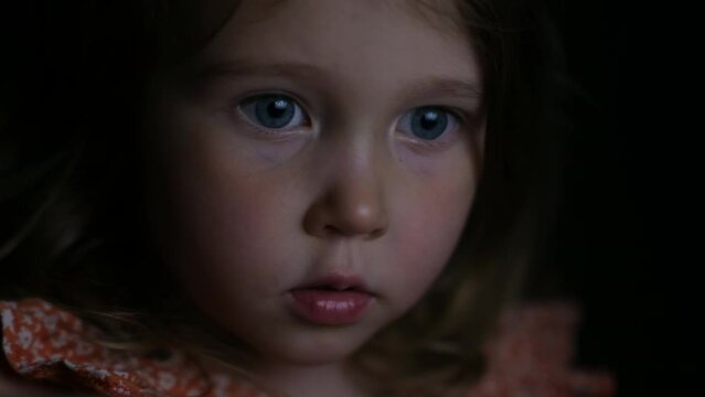 A girl of four years old is watching a cartoon on a laptop or tablet in a dark room. The light from the screen plays on the face of a beautiful girl.