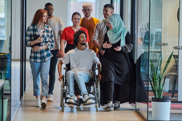 Fototapeta premium A diverse group of young business people walking a corridor in the glass-enclosed office of a modern startup, including a person in a wheelchair and a woman wearing a hijab, showing a dynamic mix of