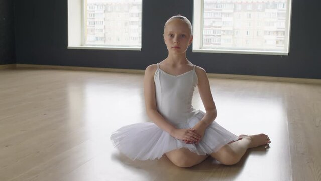 Portrait shot of little ballet school dancer sitting on floor of studio and looking at camera