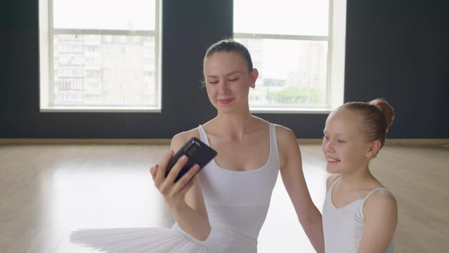 Young trainer and her small student taking selfie on smartphone while sitting on floor during break in ballet school