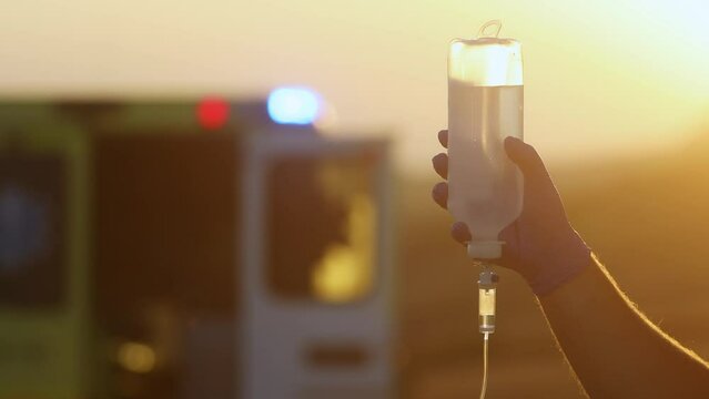Close-up hand of paramedic holding infusion set against ambulance car of emergency medical service. Themes rescue, urgency and health care..